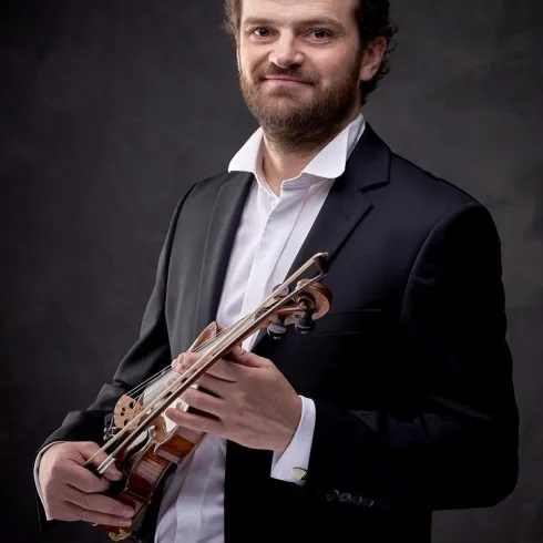 Ein Mann in einem Anzug hält eine Geige in der Hand. Der Hintergrund ist dunkel und unaufdringlich.