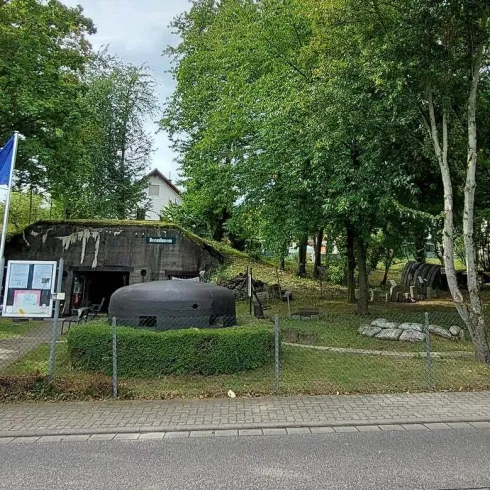 Eine kleine Anlage mit mehreren Gebäuden und einem großen Wasserbehälter in einer bewaldeten Umgebung. Im Vordergrund steht ein rotes Häuschen mit einer Flagge.