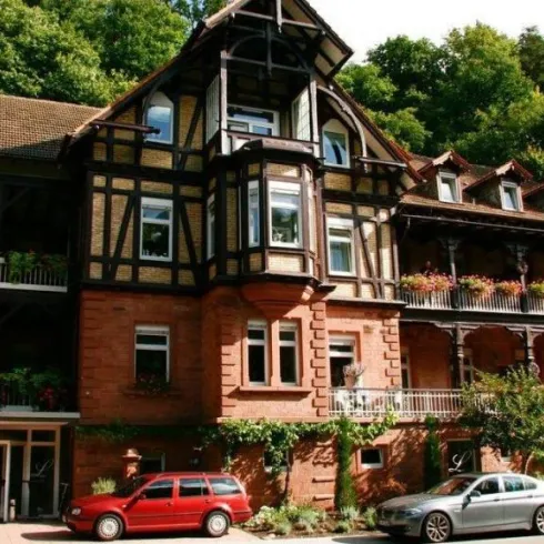 Ein charmantes Gebäude im traditionellen Stil, umgeben von Bäumen. Auf den Balkonen blühen Pflanzen, und vor dem Haus stehen einige Autos.