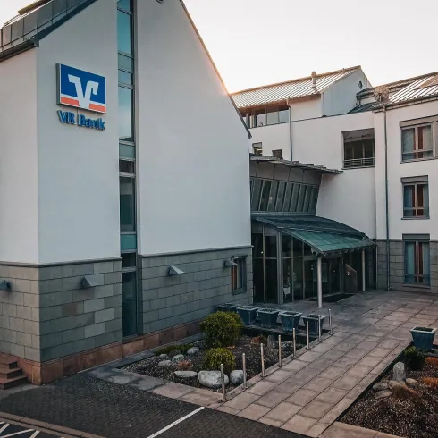 Ein modernes Gebäude mit einer Glasfront und einem klaren Design. Vor dem Haus befindet sich eine kleine Skulptur und eine gepflegte Außenfläche.