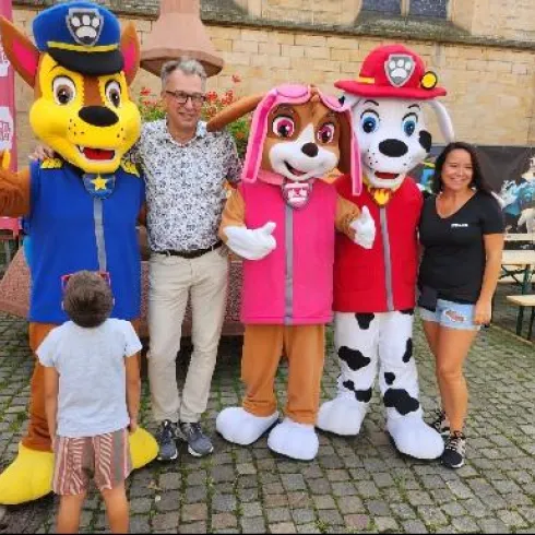 Eine Gruppe von vier Personen steht zusammen, darunter zwei Erwachsene und zwei Kinder. Die Erwachsenen sind in bunten Kostümen von Kindheitshelden bekleidet, umgeben von einer fröhlichen Atmosphäre.