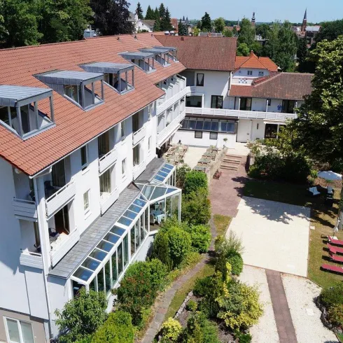 Ein modernes Hotelgebäude mit einem großen Garten. Im Vordergrund sind gemütliche Sitzgelegenheiten und gepflegte Pflanzen zu sehen.