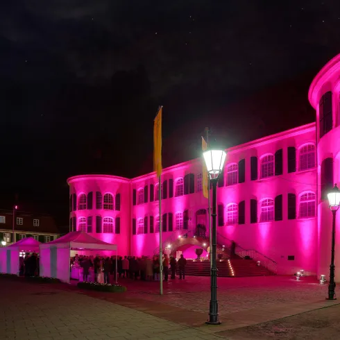 Ein historisches Gebäude, das abends pink beleuchtet ist. Im Vordergrund stehen Laternen und eine Zeltstruktur.