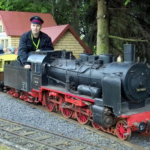 Eine kleine Dampflok fährt auf einer Modellbahnanlage. Im Hintergrund steht ein Station mit einem Lokführer.