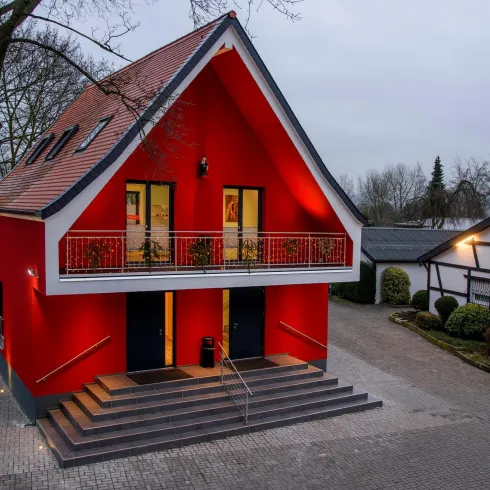Ein modernes rotes Haus mit einem schrägen Dach und großen Fenstern. Es steht auf einer gepflasterten Fläche mit Bäumen in der Nähe.