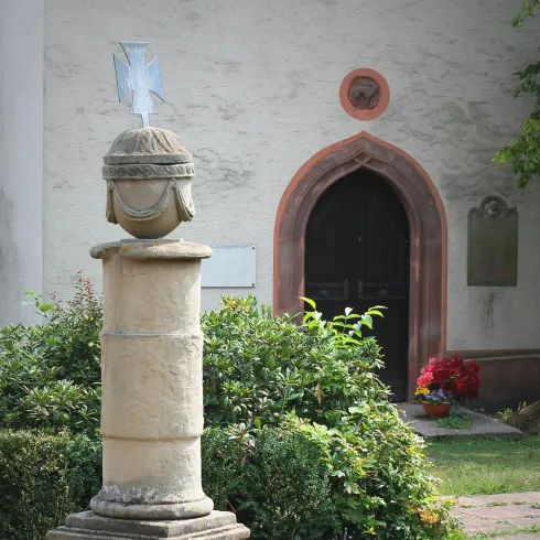 Eine kleine, historische Kirche mit steiniger Fassade und einem schrägen Dach. Im Hintergrund ist ein weiterer Kirchturm zu sehen, umgeben von Bäumen und einer ländlichen Landschaft.