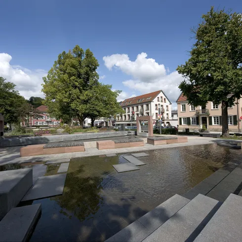 Ein attraktiver Platz mit Wasserbecken und modernen Steinbänken. Im Hintergrund sind Bäume und Gebäude zu sehen.