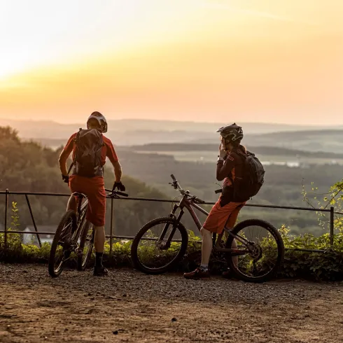 Zwei Mountainbiker stehen an einem Aussichtspunkt und genießen den Sonnenuntergang. Die Landschaft im Hintergrund zeigt sanfte Hügel und einen klaren Himmel.