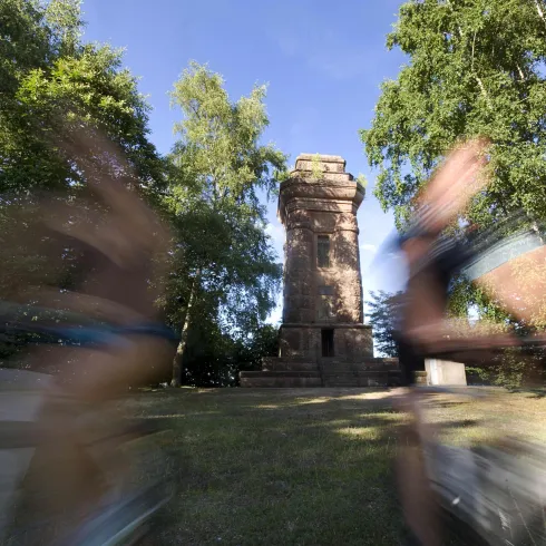 Ein historischer Turm umgeben von Bäumen. Im Vordergrund sind verschwommene Radfahrer zu sehen, die vorbeifahren.