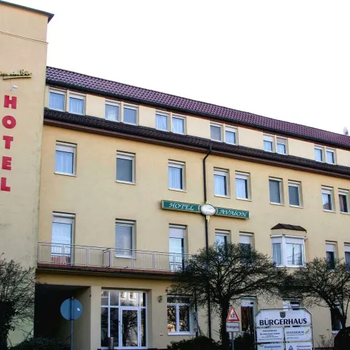 Ein Hotelgebäude mit gelber Fassade und roten Buchstaben. Vor dem Hotel befinden sich einige Bäume und ein Schild mit dem Namen des Hotels.