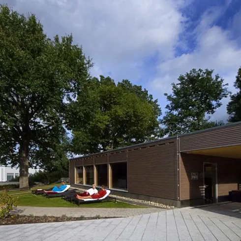 Ein modernes Gebäude mit großen Fenstern und einer Terrasse. Es gibt gemütliche Liegen im Außenbereich und viel Grün um das Haus herum.