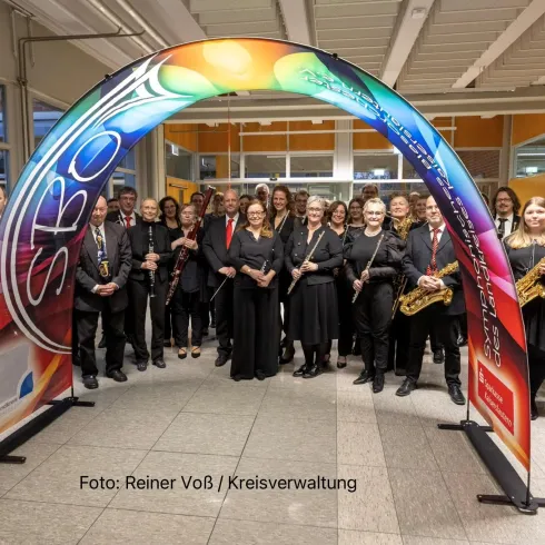 Eine große Gruppe von Musikern steht unter einem bunten Bogen. Sie tragen schwarze Kleidung und halten verschiedene Instrumente in den Händen.