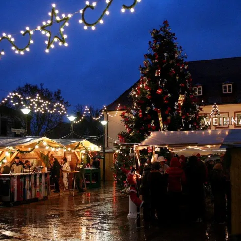 Ein Weihnachtsmarkt bei Abenddämmerung mit festlich beleuchteten Ständen und einem großen geschmückten Baum. Die Atmosphäre ist gemütlich und einladend.