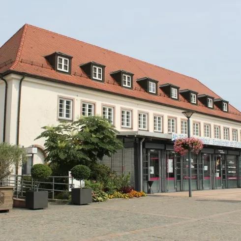 Ein modernes Gebäude mit einem roten Satteldach und großen Fenstern. Vor dem Haus gibt es Pflanzen und Blumen, die den Platz verschönern.