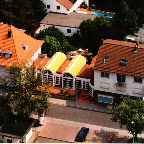 Eine Luftaufnahme von mehreren Wohngebäuden mit roten Dächern. Im Vordergrund befinden sich hellgelbe Gewächshäuser und ein gepflegter Garten.