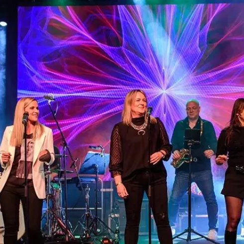 Eine Liveband steht auf der Bühne und spielt Musik. Bunte Lichter und eine dynamische Hintergrundprojektion schaffen eine fröhliche Atmosphäre.