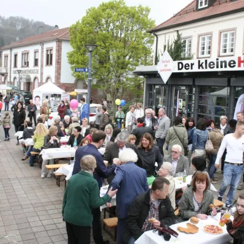 Eine belebte Straße mit vielen Menschen, die an Tischen sitzen und essen. Einige Gäste feiern mit Luftballons und eine freundliche Atmosphäre ist zu spüren.