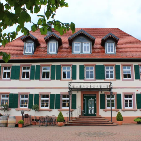 Ein schönes Gebäude mit einem roten Dach und grünen Fensterläden. Der Eingangsbereich ist freundlich gestaltet und von Pflanzen umgeben.