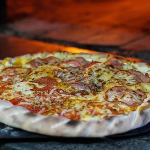 Eine frisch gebackene Pizza mit Käse und Schinken. Sie liegt auf einem schwarzen Blech vor dem Hintergrund eines warmen Backofens.