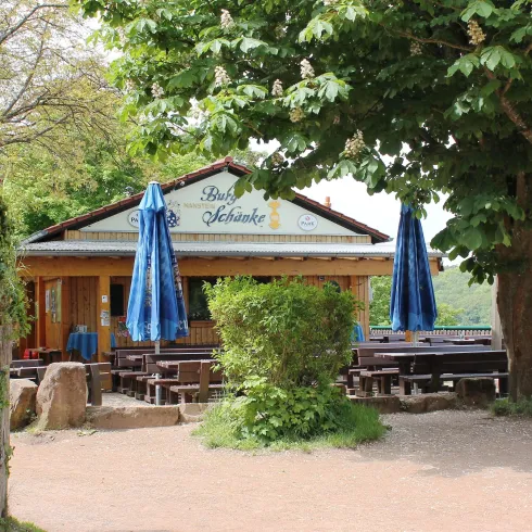 Ein einladendes Gasthaus mit blauen Sonnenschirmen und einem schönen Garten. Im Hintergrund sind Bäume und eine grüne Landschaft zu sehen.