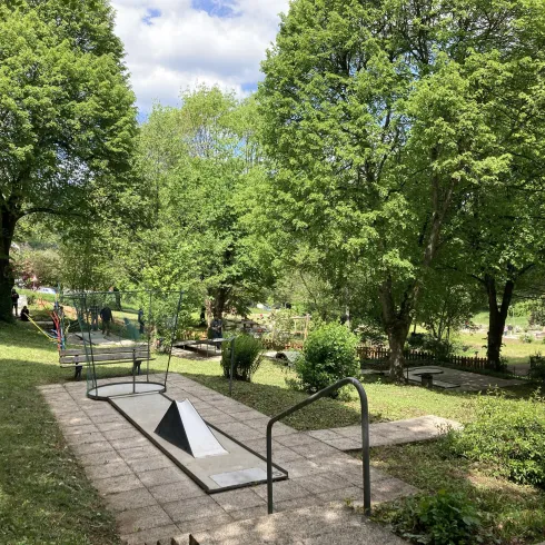 Eine grüne Parklandschaft mit vielen Bäumen und einer gepflasterten Fläche. Im Hintergrund sind weitere Grünflächen und Spaziergänger zu sehen.