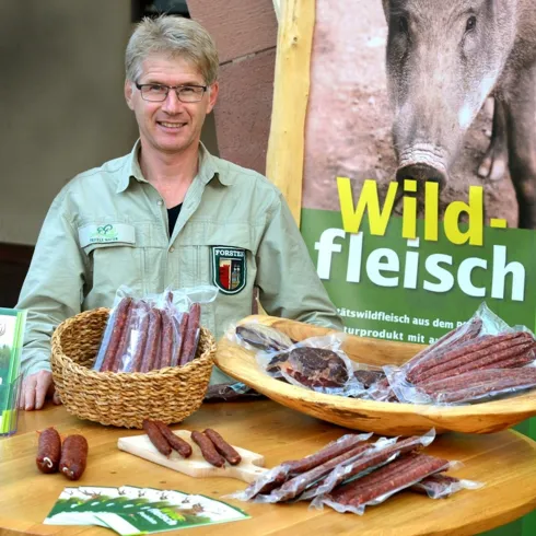 Ein Mann steht an einem Tisch mit verschiedenen Wildfleischprodukten. Im Hintergrund ist ein Banner mit der Aufschrift „Wildfleisch“ zu sehen.