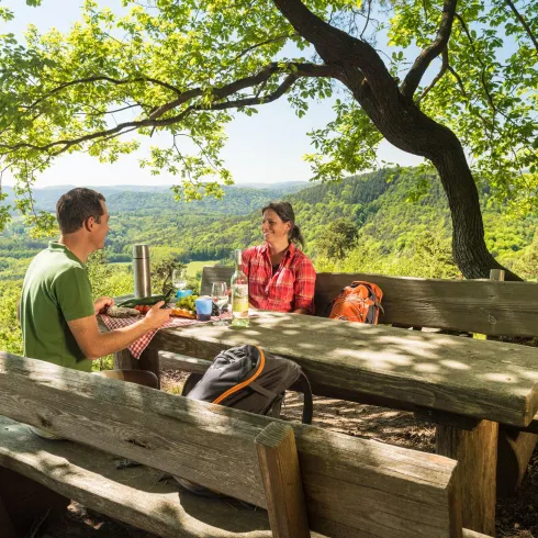 Ein Tisch in der Natur, an dem zwei Personen eine Pause machen. Im Hintergrund sieht man eine wunderschöne Landschaft mit Bäumen und Hügeln.