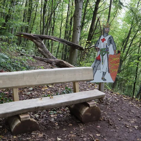 Eine hölzerne Bank steht in einem Wald. Neben der Bank steht eine Figuren-Darstellung eines Ritters.