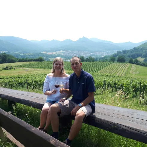 Ein Paar sitzt auf einer Bank und genießt ein Getränk inmitten von Weinbergen. Im Hintergrund sind Hügel und eine malerische Landschaft zu sehen.
