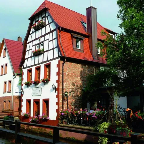 Ein charmantes, historisches Gebäude mit einem roten Dach und Fachwerk an einer malerischen Straße. Bunte Blumen schmücken die Fenster und den Eingangsbereich.