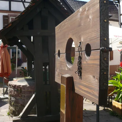 Eine historische Holzstange mit zwei Löchern steht in einer Altstadt. Im Hintergrund ist ein traditionelles Gebäude sichtbar.