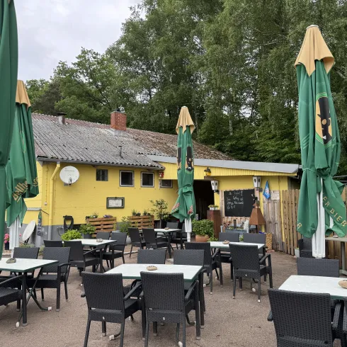 Ein gemütliches Café mit vielen Tischen und Sonnenschirmen steht vor einem gelben Gebäude. Die Umgebung ist grün mit Bäumen und bietet eine einladende Atmosphäre.