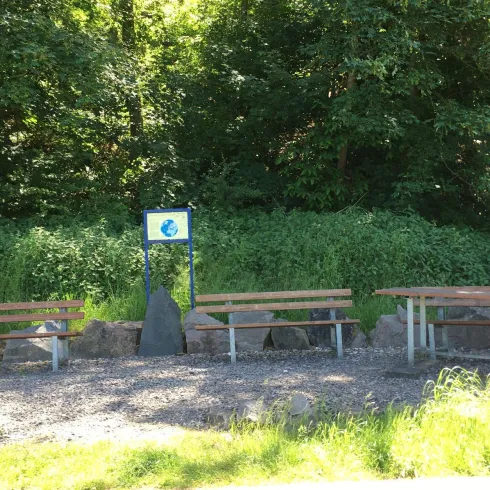 Ein Rastplatz mit mehreren Holzbänken und einem Informationsschild. Umgeben von grünen Bäumen und Sträuchern.