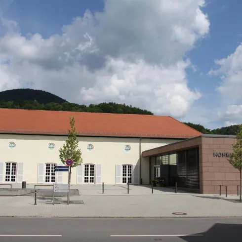 Ein modernes Gebäude mit einem roten Dach und großen Fenstern. Im Hintergrund sieht man hügelige Landschaften und einen klaren Himmel.