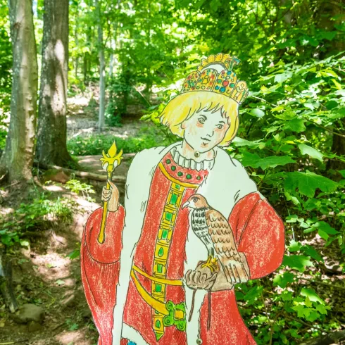 Eine farbenfrohe Pappfigur eines Königs steht im Wald. Der König hält einen Vogel und trägt eine Krone sowie prächtige Gewänder.