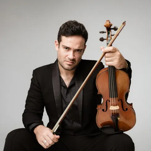 Ein Musiker sitzt auf dem Boden und hält eine Geige in der Hand. Er trägt einen eleganten schwarzen Anzug und schaut direkt in die Kamera.