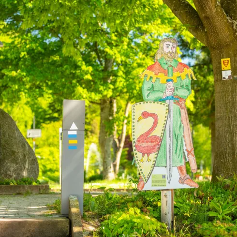 Ein aufgestellter Ritter mit Schwert und Schild steht in einem Park. Im Hintergrund sind grüne Bäume und eine Gedenktafel sichtbar.