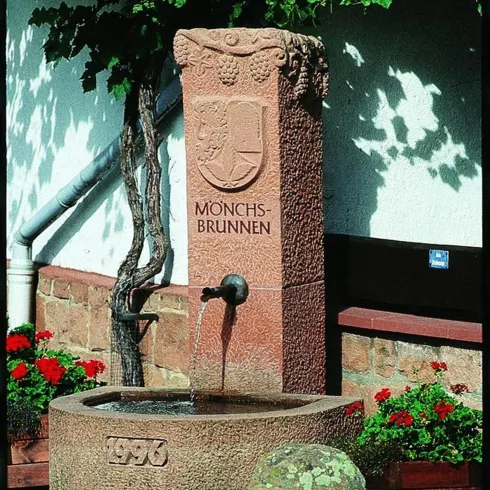 Ein schöner Brunnen aus Stein mit der Aufschrift "Mönchs-Brunnen" und dem Jahr 1996. Umgeben von blühenden Blumen und einer grünen Landschaft.