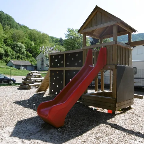 Ein Spielplatz mit einer roten Rutsche und einem Holzturm. Im Hintergrund sind Grünflächen und einige Wohnmobile zu sehen.