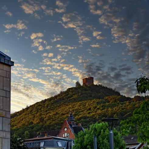 Ein Hügel mit einer Burgruine steht vor einem Himmel mit vielen Wolken. Im Vordergrund sind moderne Gebäude und Bäume sichtbar.