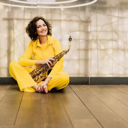 Eine lächelnde Frau sitzt auf dem Boden und hält ein Saxophon in der Hand. Sie trägt einen gelben Anzug und hat lockige Haare.