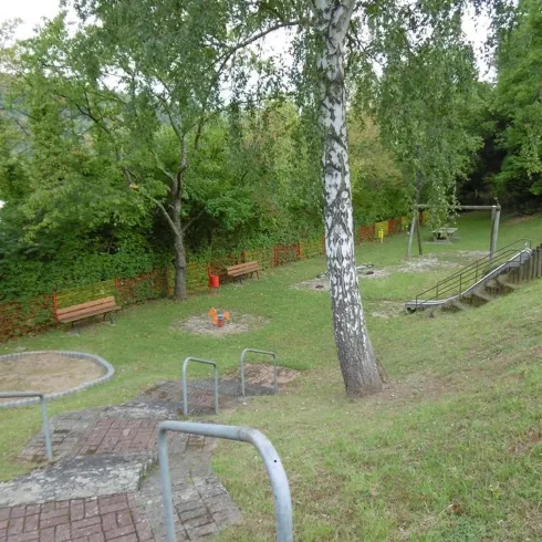 Ein kleiner Park mit Bänken, Spielgeräten und grüner Vegetation. Der Bereich hat eine leichte Hanglage und mehrere Spielmöglichkeiten.