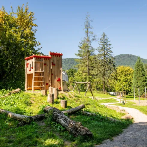 Ein Spielplatz mit einem Holzspielhaus und Schaukeln in einer grünen Parklandschaft. Im Hintergrund sind Bäume und Hügel sichtbar.