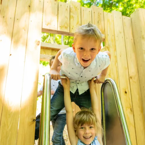 Zwei Kinder spielen auf einer Rutsche in einem Spielplatz. Die Sonne scheint durch die Bäume und sorgt für eine fröhliche Atmosphäre.