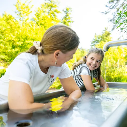 Eine Frau und ein Mädchen genießen entspannte Zeit in einem Außenbecken. Umgeben von grünem Gras und Bäumen strahlt die Szene Freude und Erholung aus.