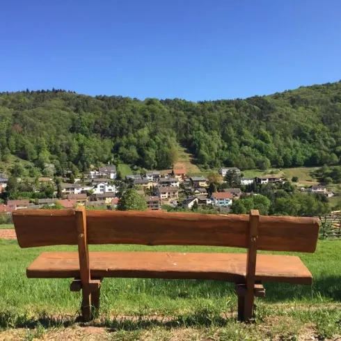 Eine Holzbank steht auf einer Wiese mit Blick auf ein Dorf und grüne Hügel. Der Himmel ist klar und blau.