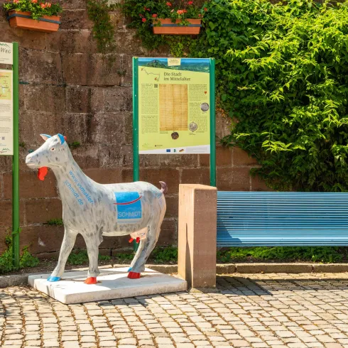 Eine bunte Ziegenskulptur steht neben einem blauen Sitzbank. Im Hintergrund sind Informationsschilder angebracht.