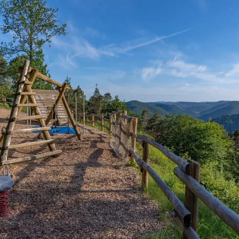 Ein Spielplatz mit einer Kletterstruktur und einem schönen Blick auf die umliegenden Berge. Die Szene ist umgeben von viel Grün und einem klaren Himmel.