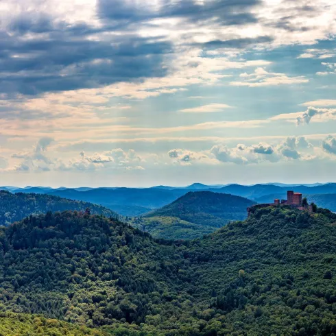 Eine Panoramasicht auf grüne Hügel unter einem teilweise bewölkten Himmel. In der Ferne sind Berge und eine Burgruine sichtbar.