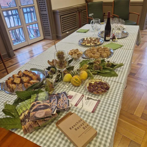 Ein gedeckter Tisch mit verschiedenen Leckereien wie Gebäck, Obst und einer Flasche Wein. Im Hintergrund sind Stühle und Fenster zu sehen.